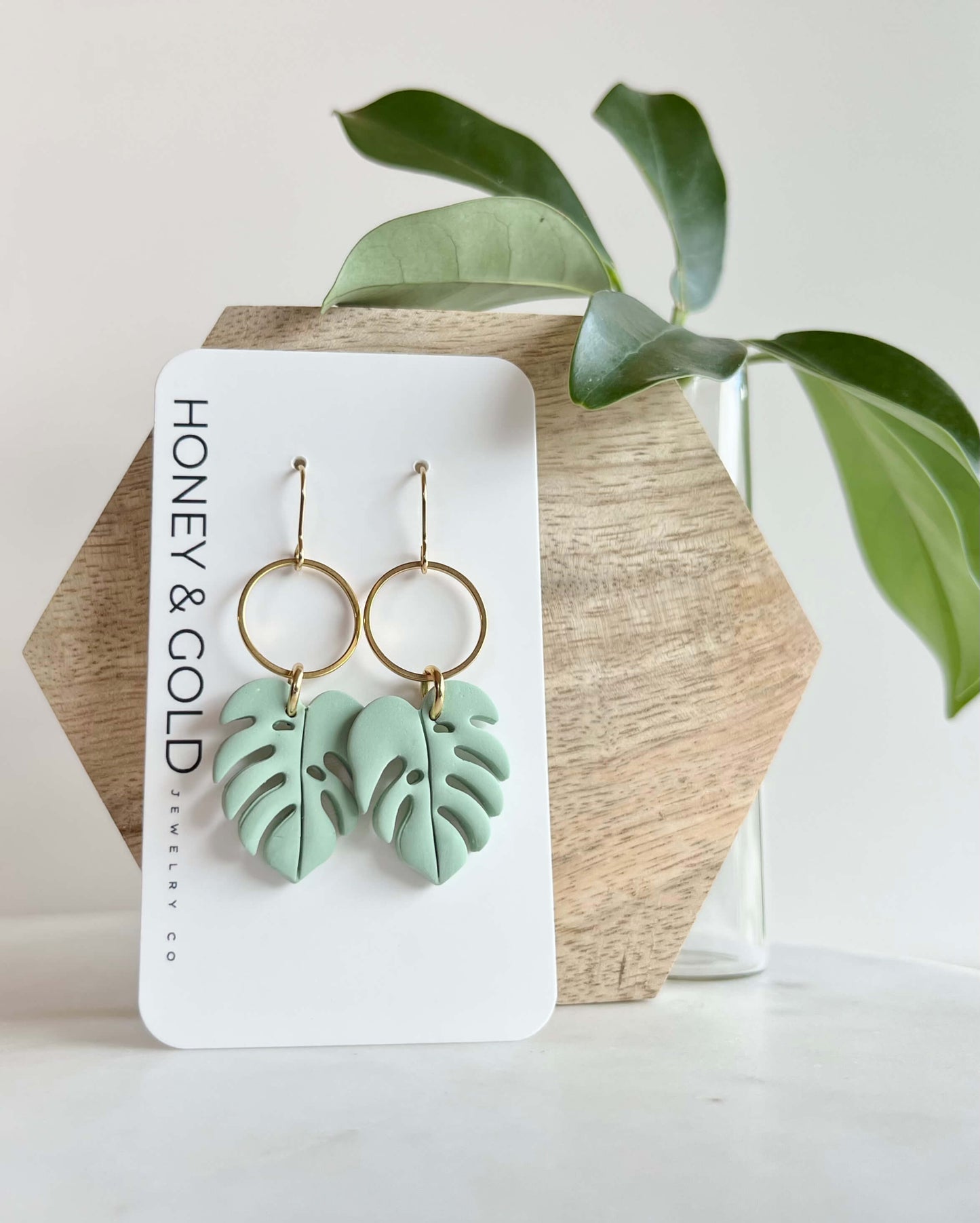Green leaf-shaped earrings on a Honey & Gold branded card with a plant in the background.
