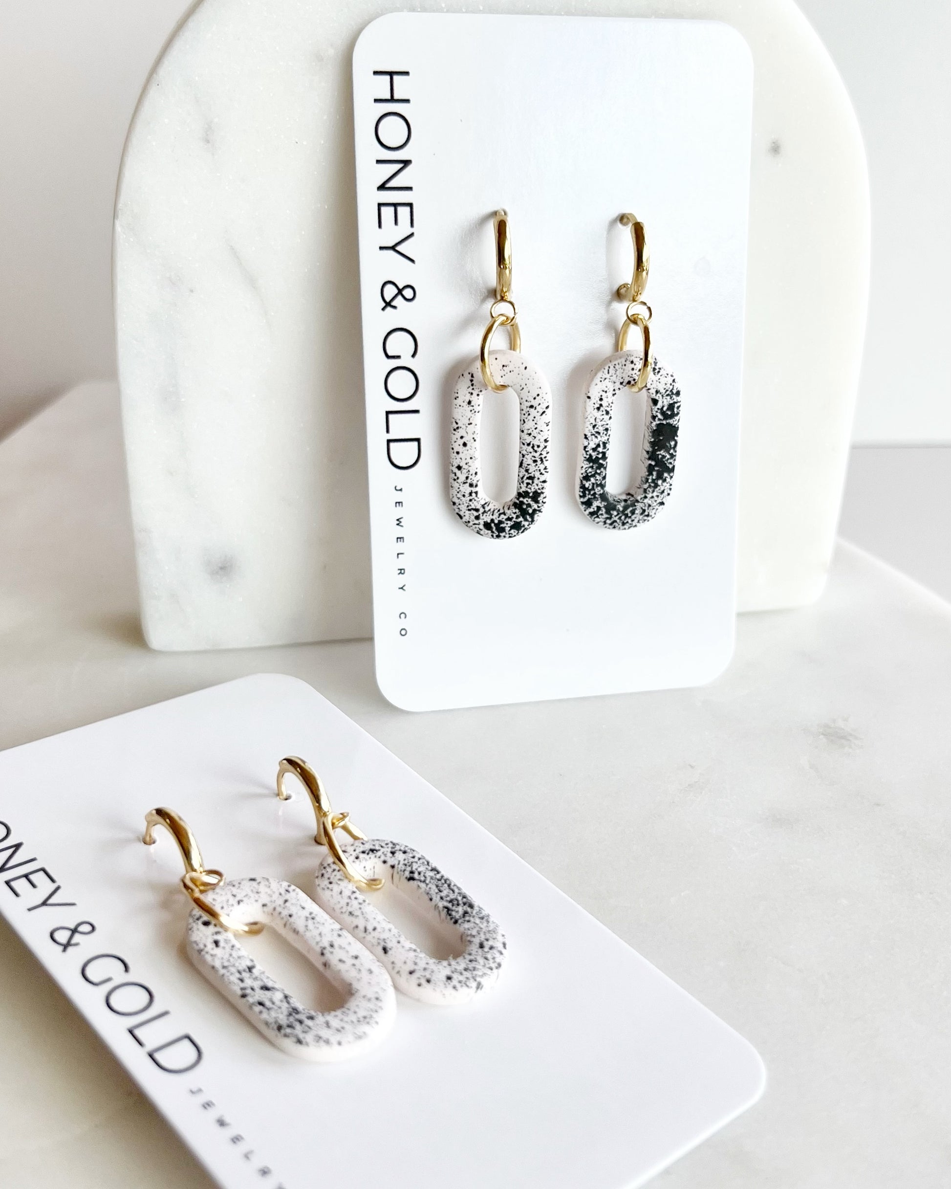 Handmade polymer clay earrings featuring black speckles on white oval links with gold-plated huggie hoops, photographed on a neutral background.

