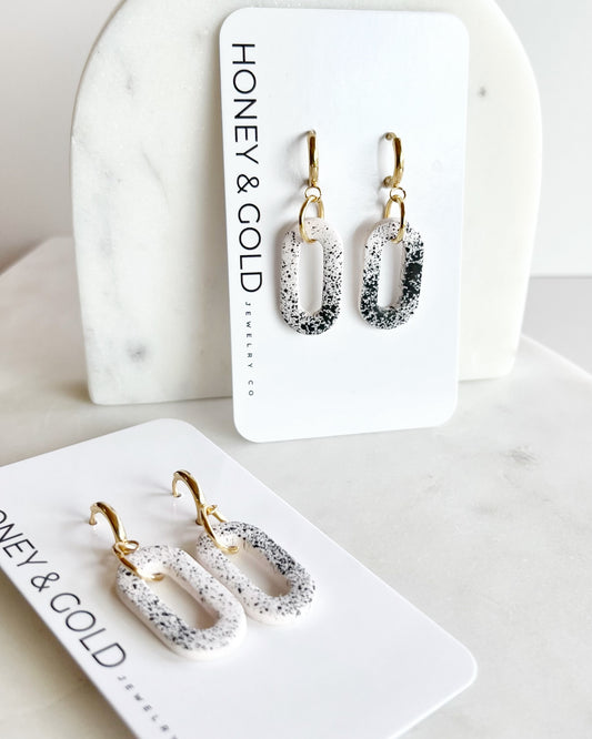 Handmade polymer clay earrings featuring black speckles on white oval links with gold-plated huggie hoops, photographed on a neutral background.

