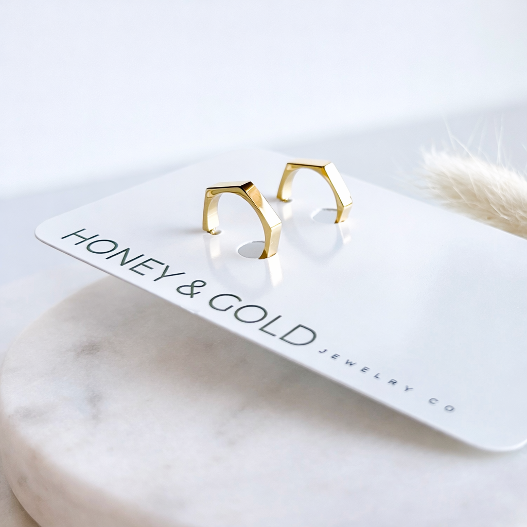 Gold geometric earrings on a 'Honey & Gold Jewelry Co' card with a marble surface background.