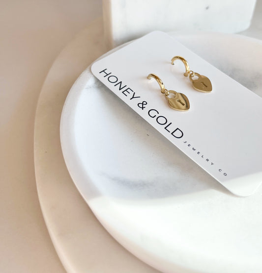 Gold heart lock huggie earrings displayed on Honey & Gold Jewelry Co. card, placed on white marble plates.