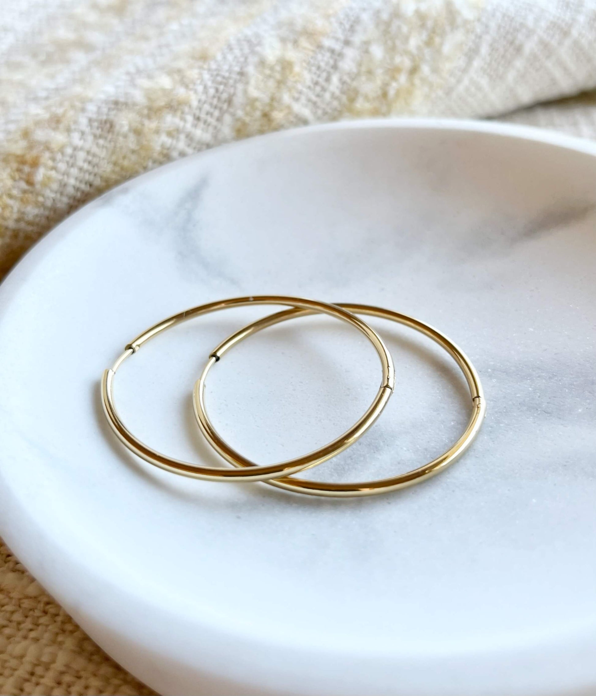18K gold hoop earrings on a soft white background with soft marbled accents. Smooth, minimalist design with a 2mm thick profile.