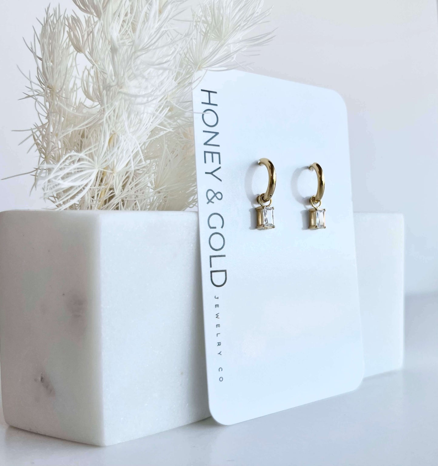 Gold huggie hoop earrings with clear rectangular zirconia charms hanging on a gold rod, styled with soft white background and minimal marble accents.