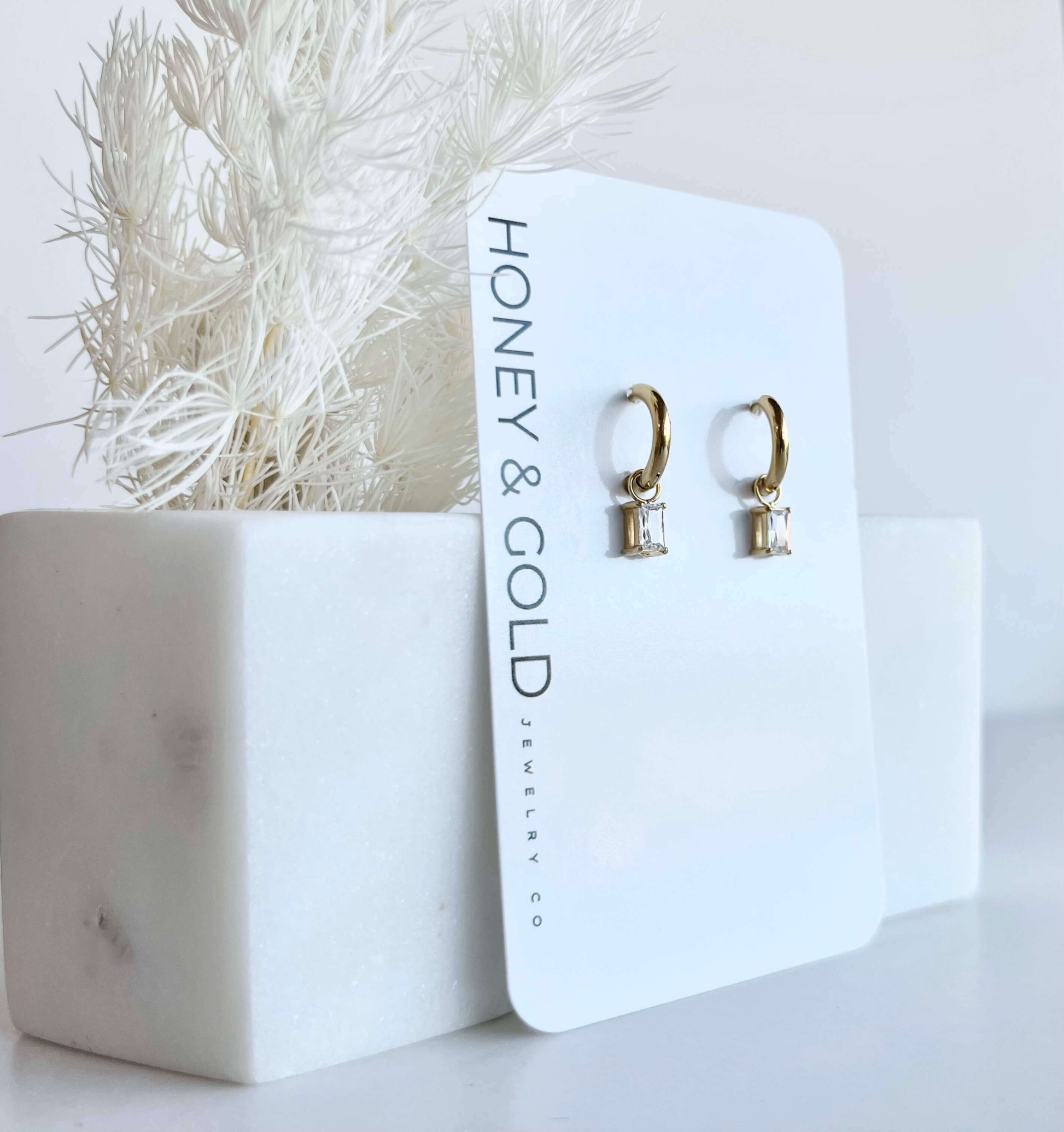 Gold huggie hoop earrings with clear rectangular zirconia charms hanging on a gold rod, styled with soft white background and minimal marble accents.