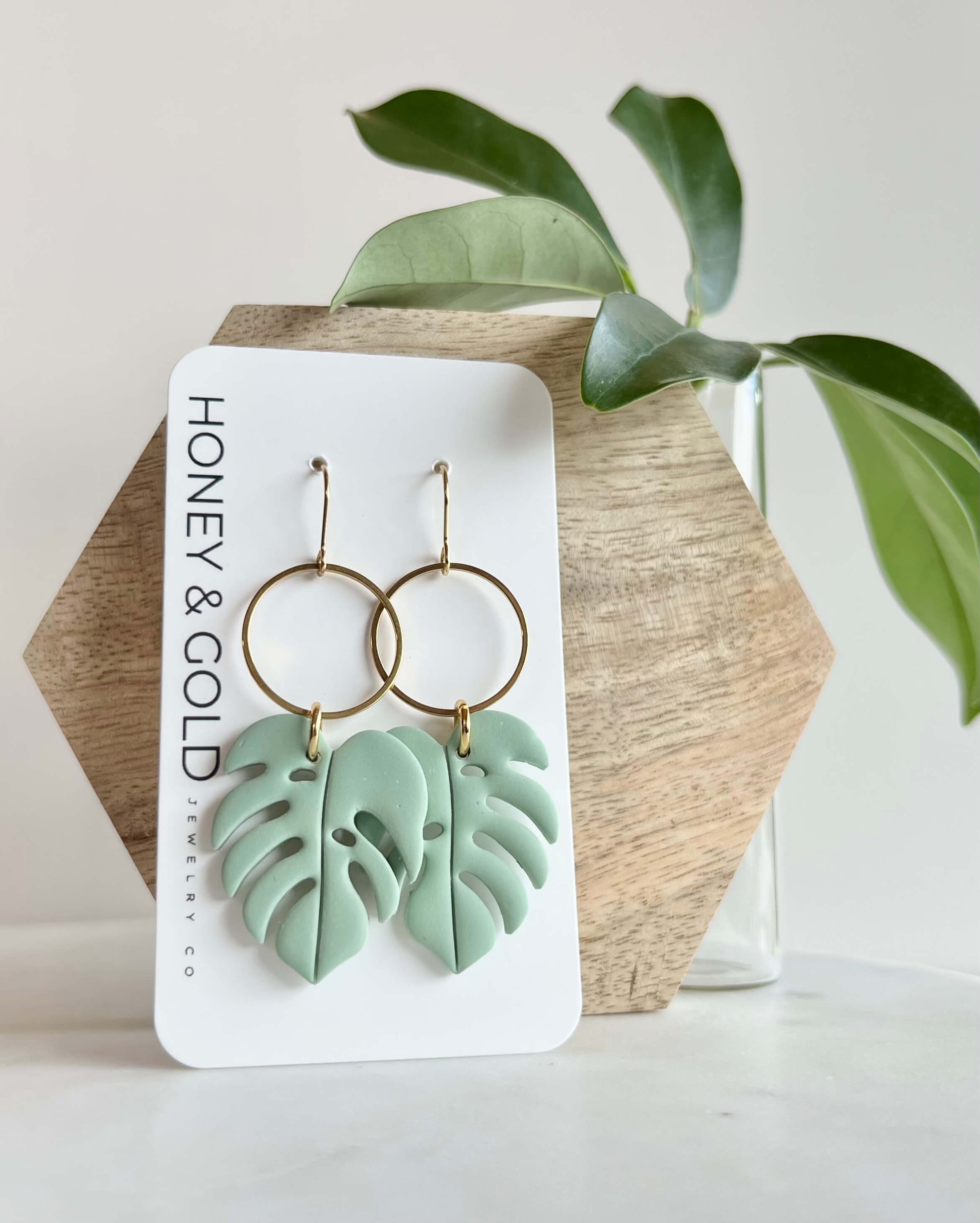 Green leaf-shaped earrings on a Honey & Gold branded card with a plant in the background