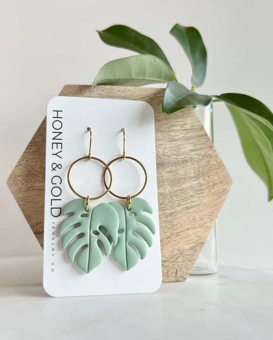 Green leaf-shaped earrings on a Honey & Gold branded card with a plant in the background