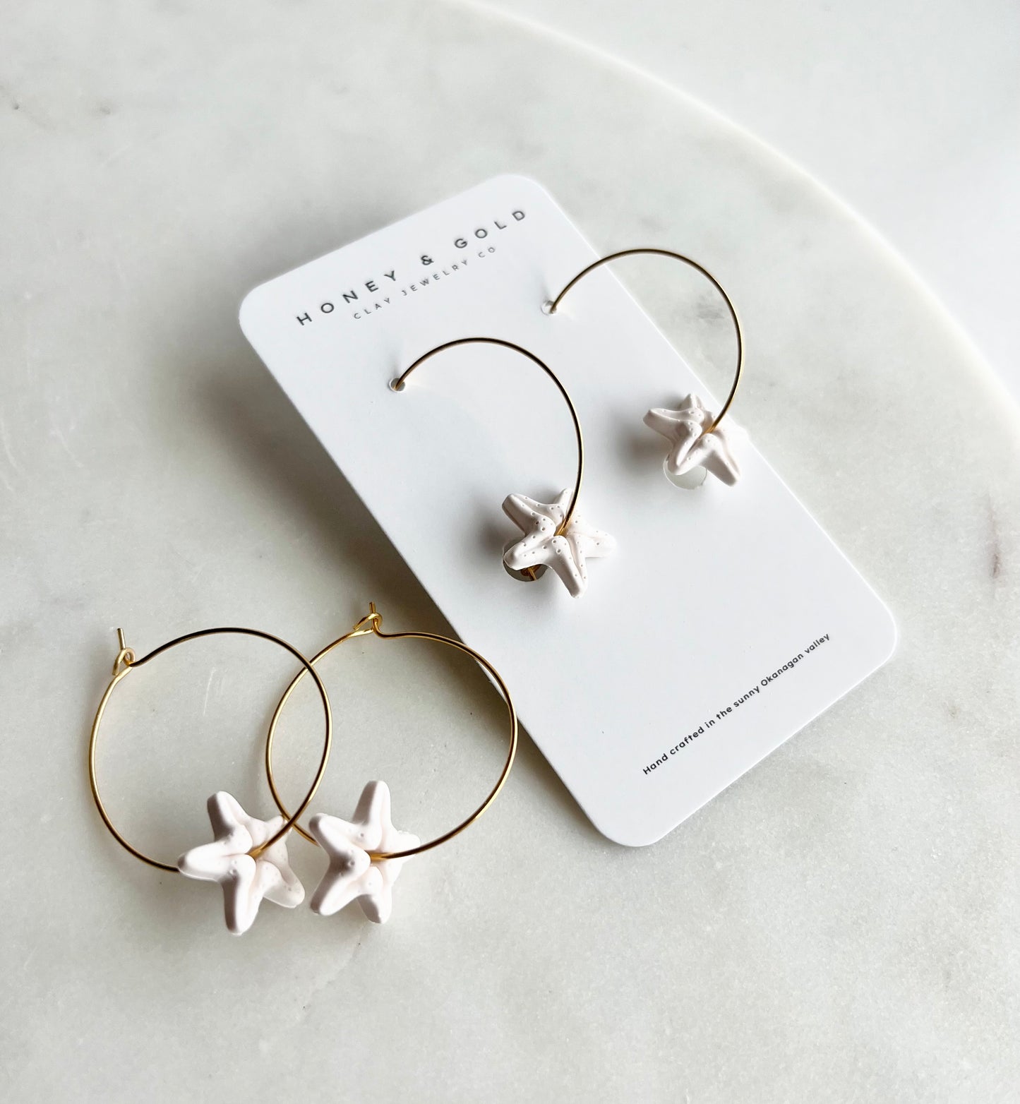 A pair of hoop earrings made from hypoallergenic stainless steel, designed for sensitive ears. The earrings feature a soft white clay starfish hanging below, creating a blend of beachy and boho aesthetics. The lightweight design offers comfort for all-day wear, and the soft white color adds a touch of coastal charm to any outfit.