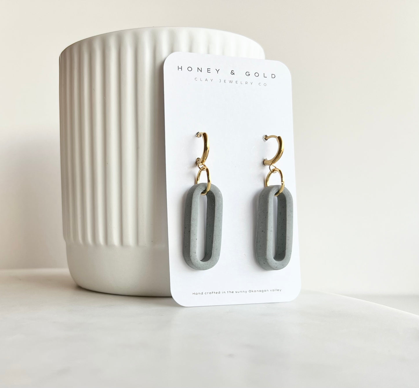 Modern earrings on a white marbled background, featuring a lightweight grey design with gold accents. The earrings have an oval shape and soft stone hues, creating an elegant and stylish appearance."